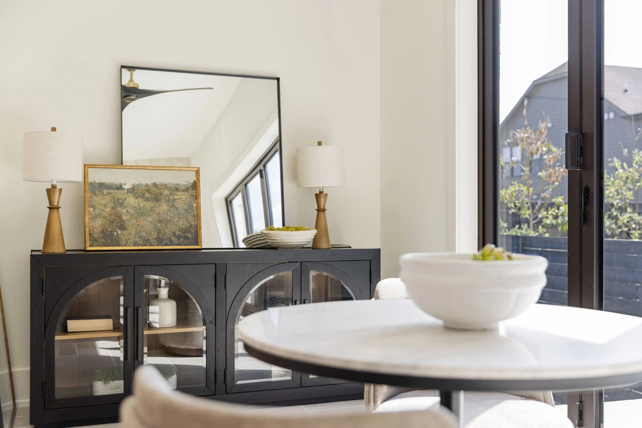 Black sideboard with arched glass doors styled with paired wood lamps, a leaning mirror, and a landscape painting beside a round marble table