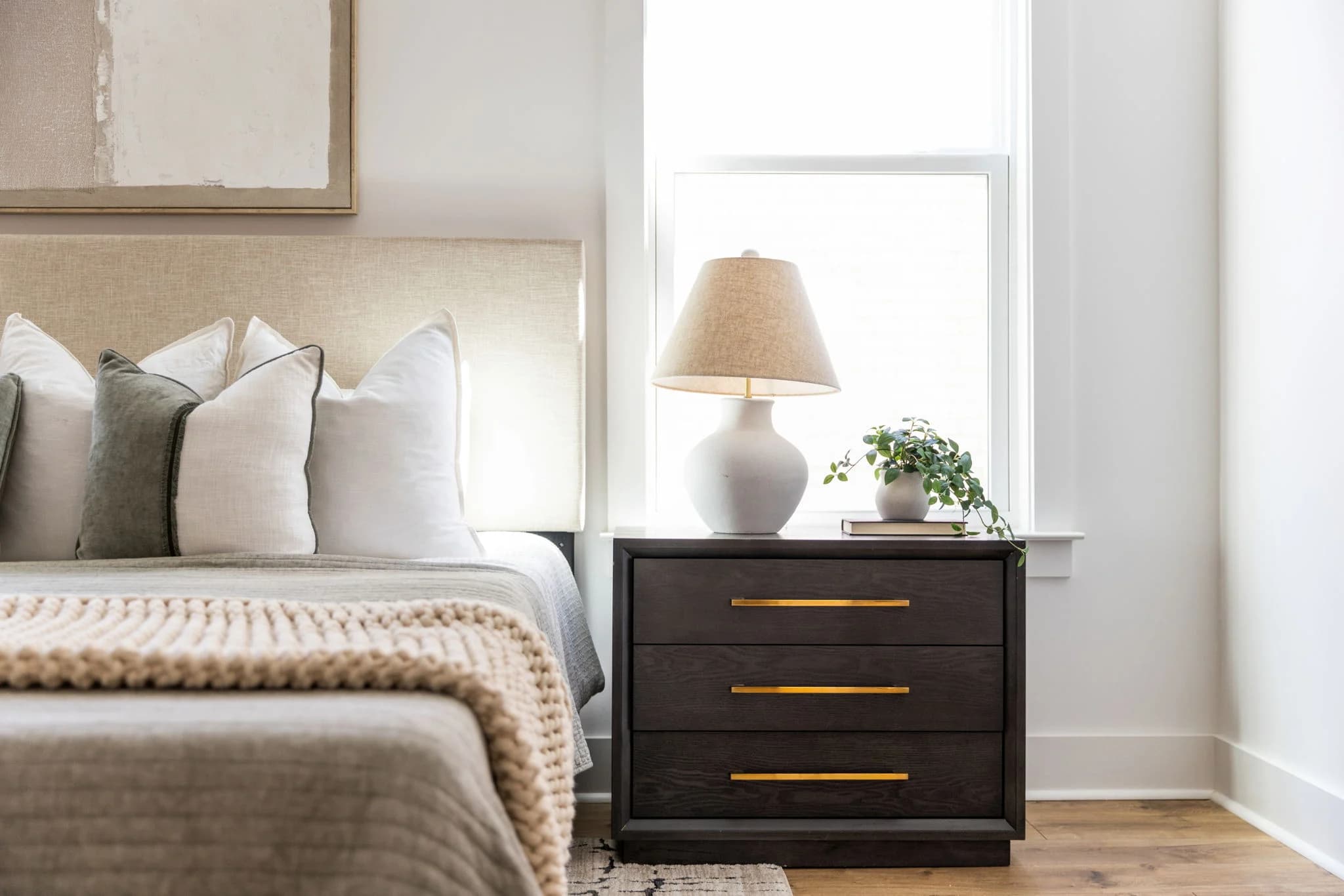 Bed with a knit throw beside a dark wood nightstand with brass pulls and a white ceramic lamp