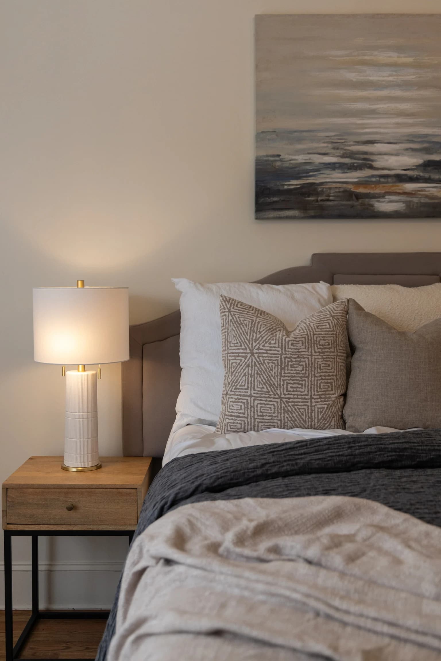 Bedside nightstand with a white lamp next to a bed with textured pillows and an ocean painting