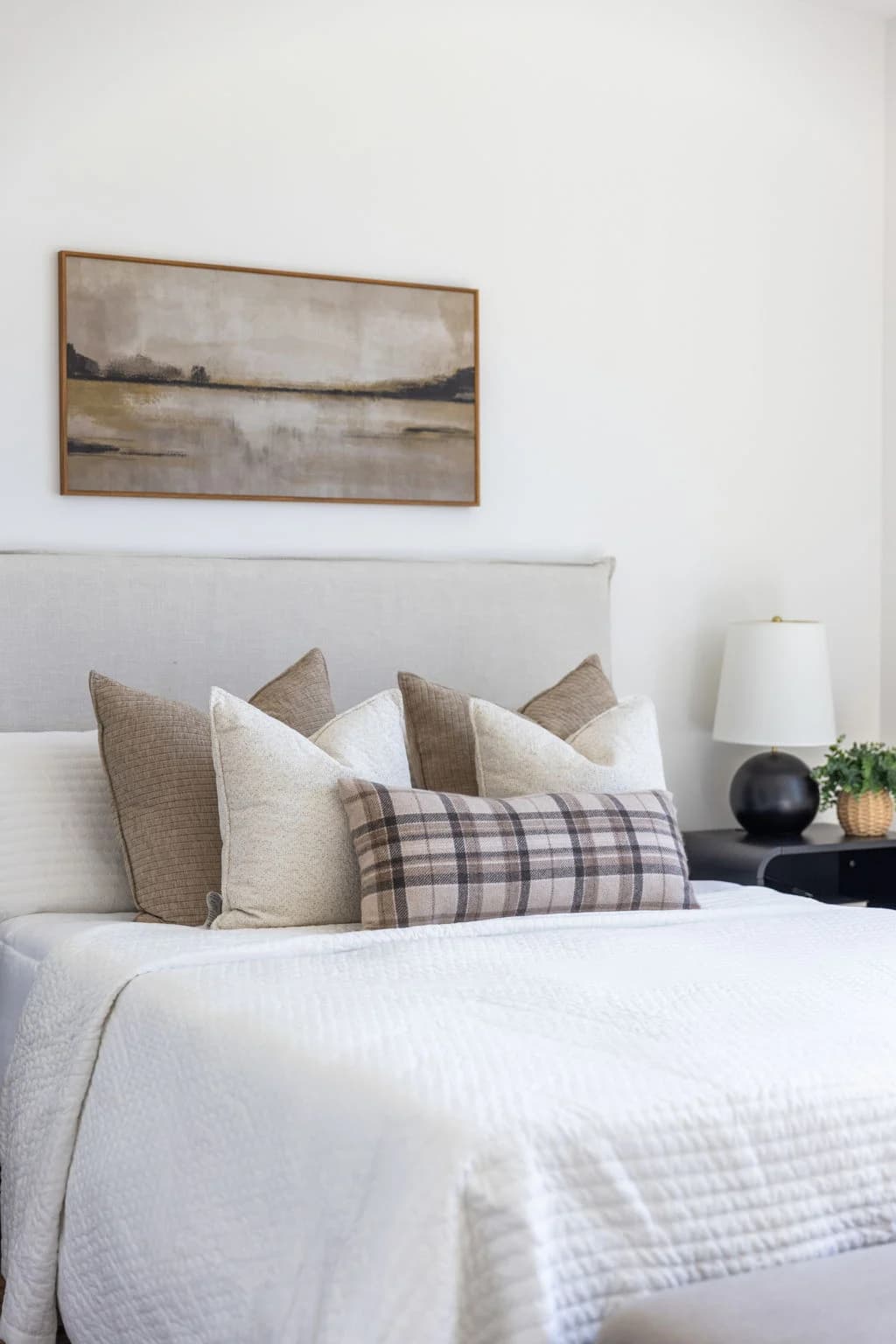 Bed layered with cream and plaid pillows beside a black lamp, below a framed landscape painting