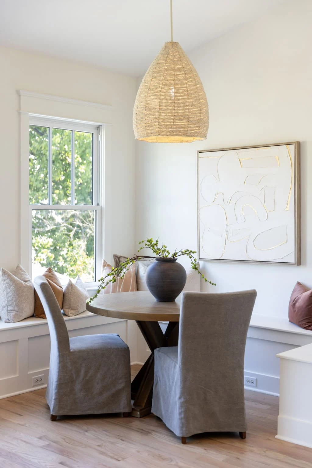 Breakfast nook with a window banquette, a round table, gray slipcovered chairs, and a woven pendant