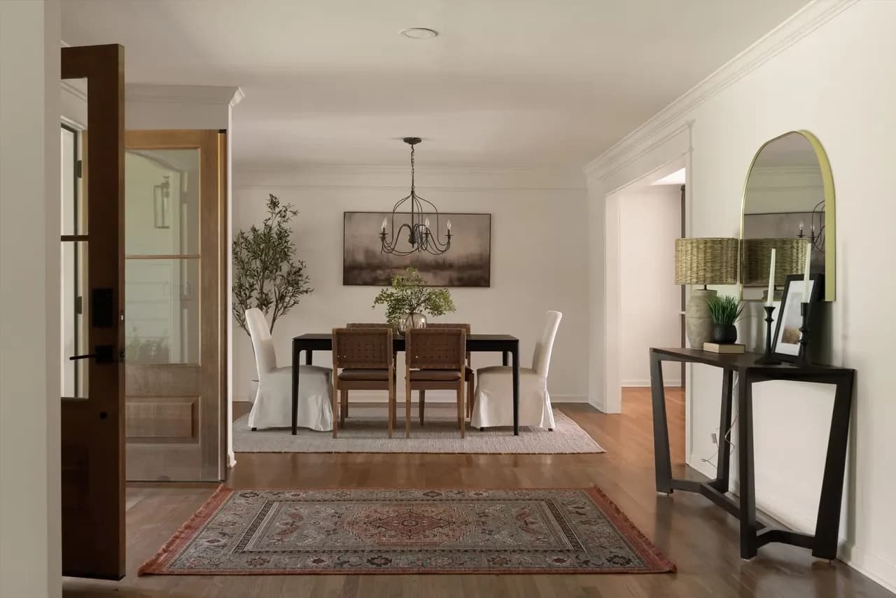 Dining room with a wood table, cream slipcovered chairs, and an iron chandelier, seen past an arched mirror over a black console