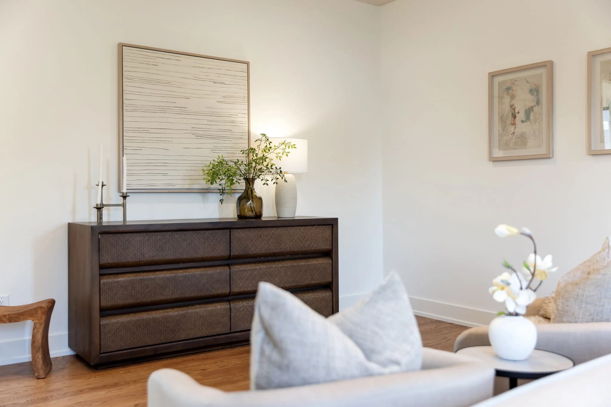 Dark wood dresser styled with a lamp, greenery, and striped art behind a cream sofa with an orchid