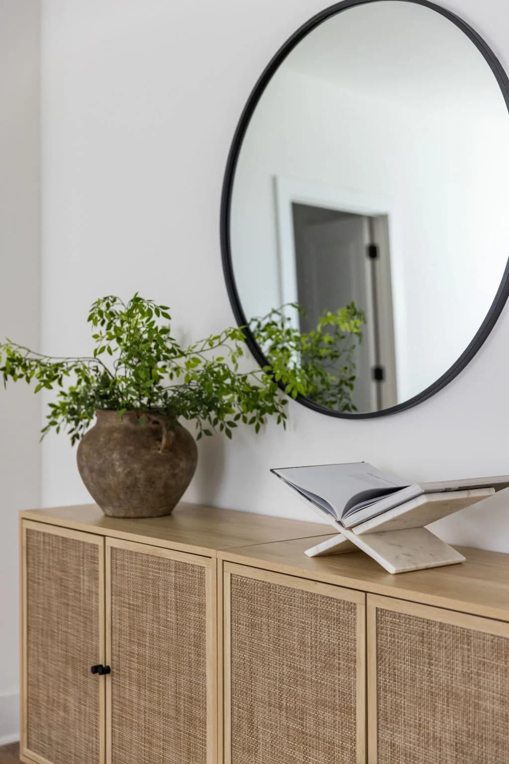 Cane front cabinet styled with a clay vase of greenery and an open book below a round black mirror