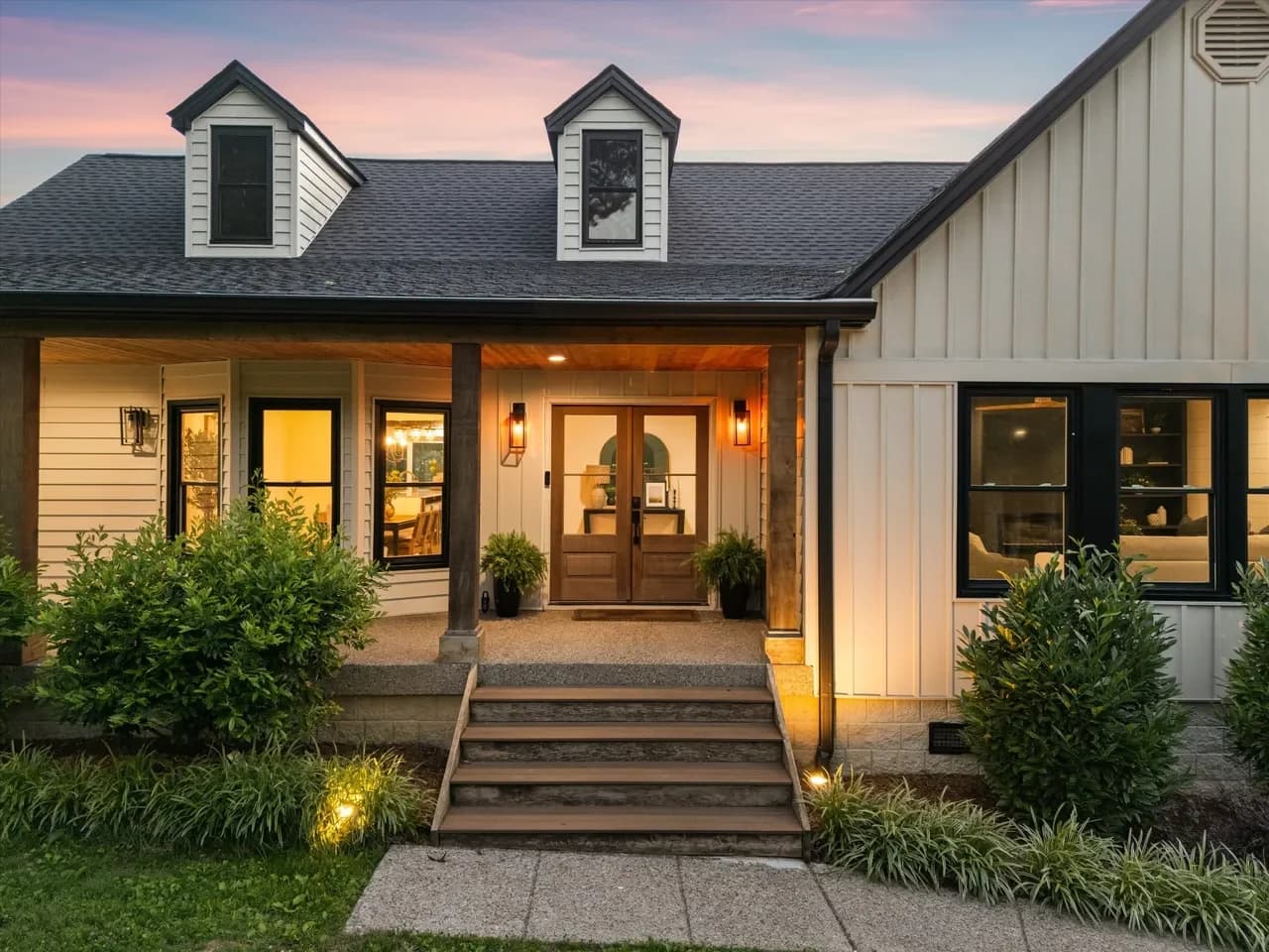 Farmhouse exterior at dusk with board and batten siding, two dormers, and a lit covered porch with double wood doors