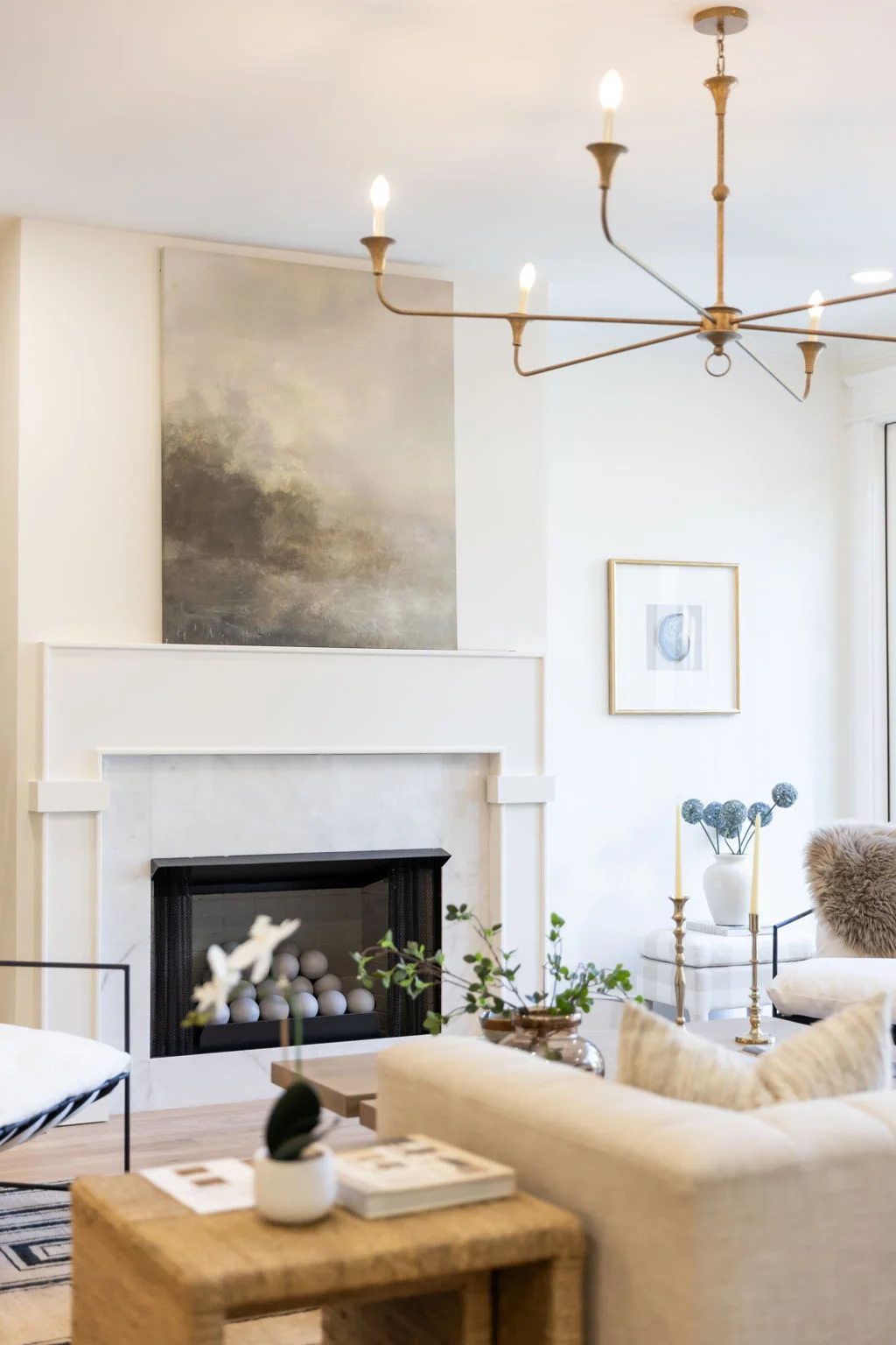 White fireplace with moody abstract art and a brass chandelier above a cream sofa and woven side table