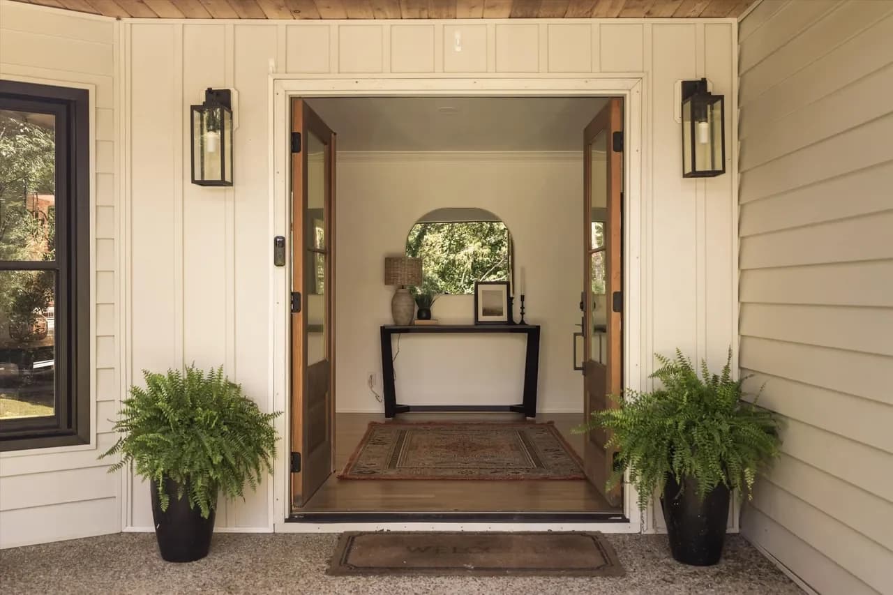 Front porch with open double wood doors to an entry with an arched mirror over a console, flanked by potted ferns
