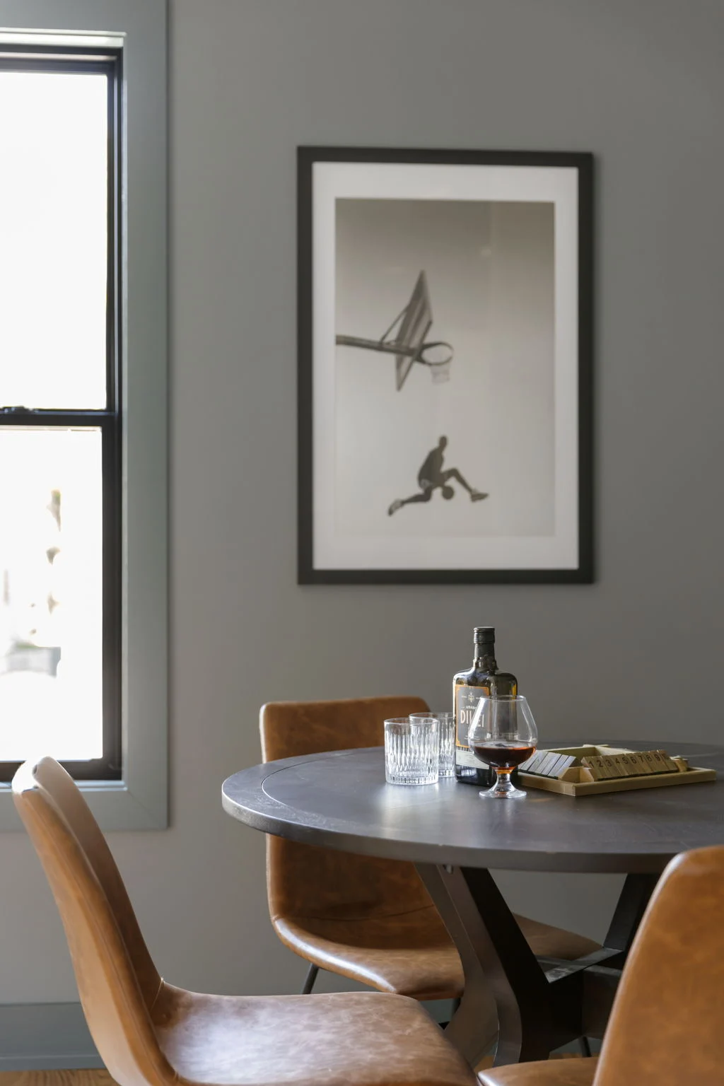Round game table with cognac leather chairs below framed black and white basketball art on a gray wall