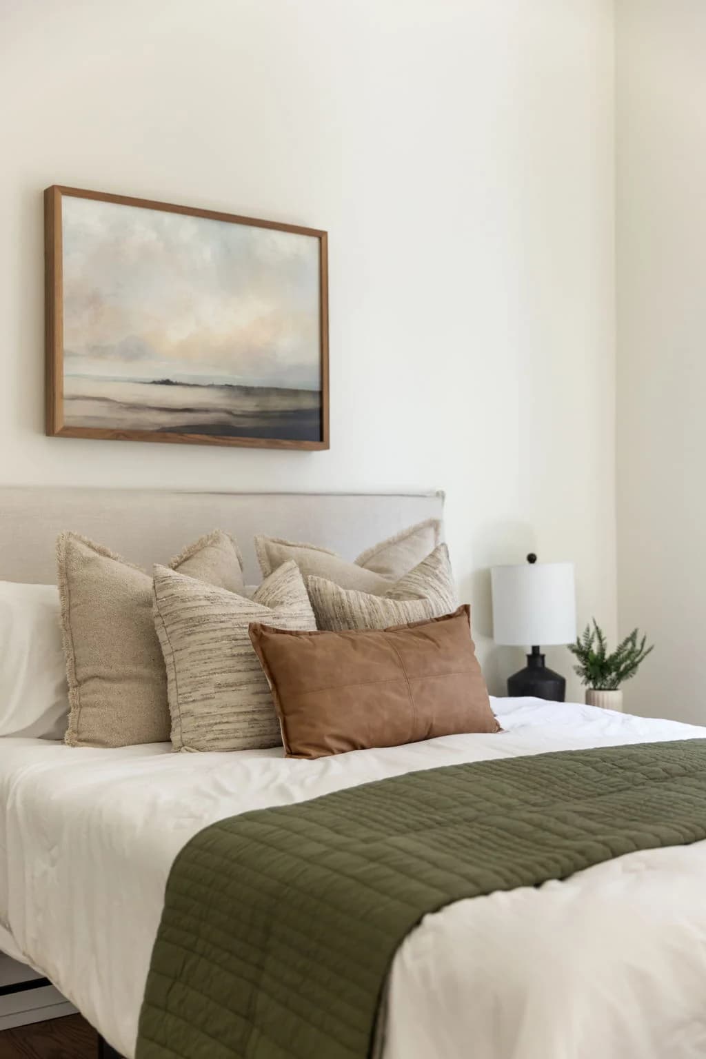 Bed with an olive green throw and layered neutral pillows below a coastal painting