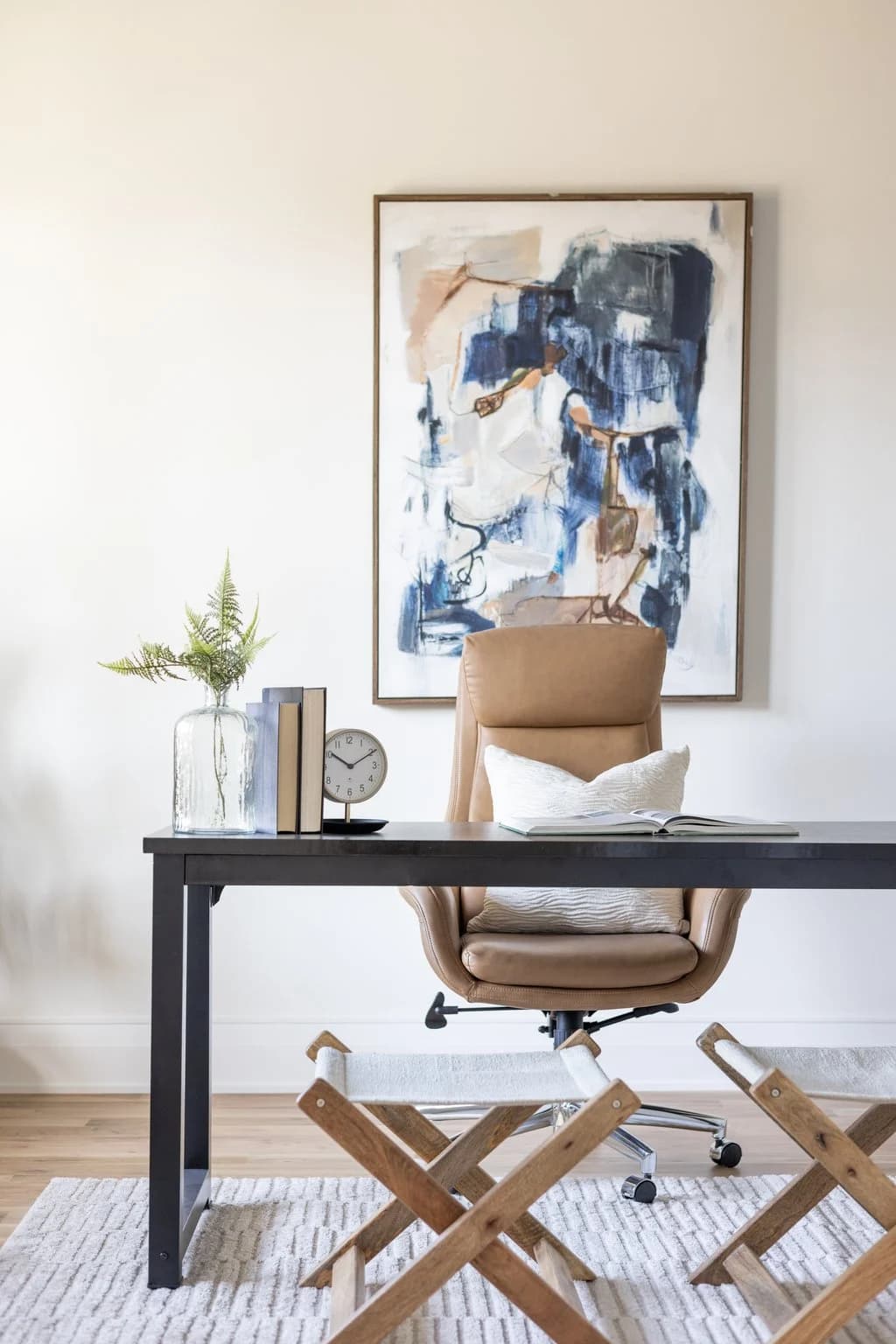 Home office with a black desk, tan leather chair, abstract blue art, and a folding X bench
