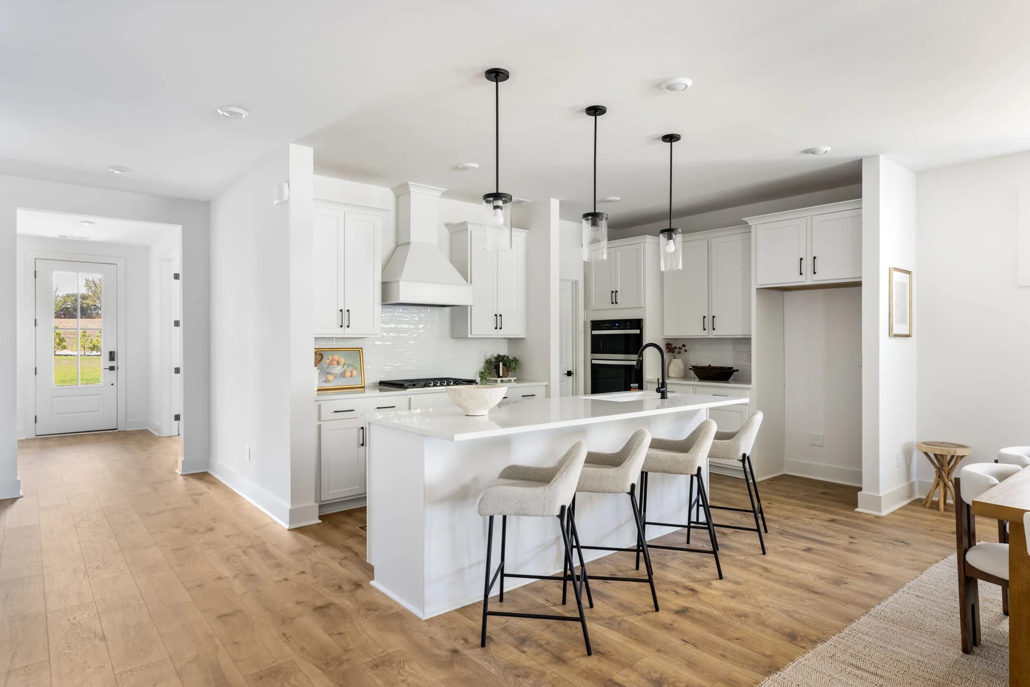 White kitchen with a long island, four cream counter stools, glass pendant lights, and a hallway to the front door