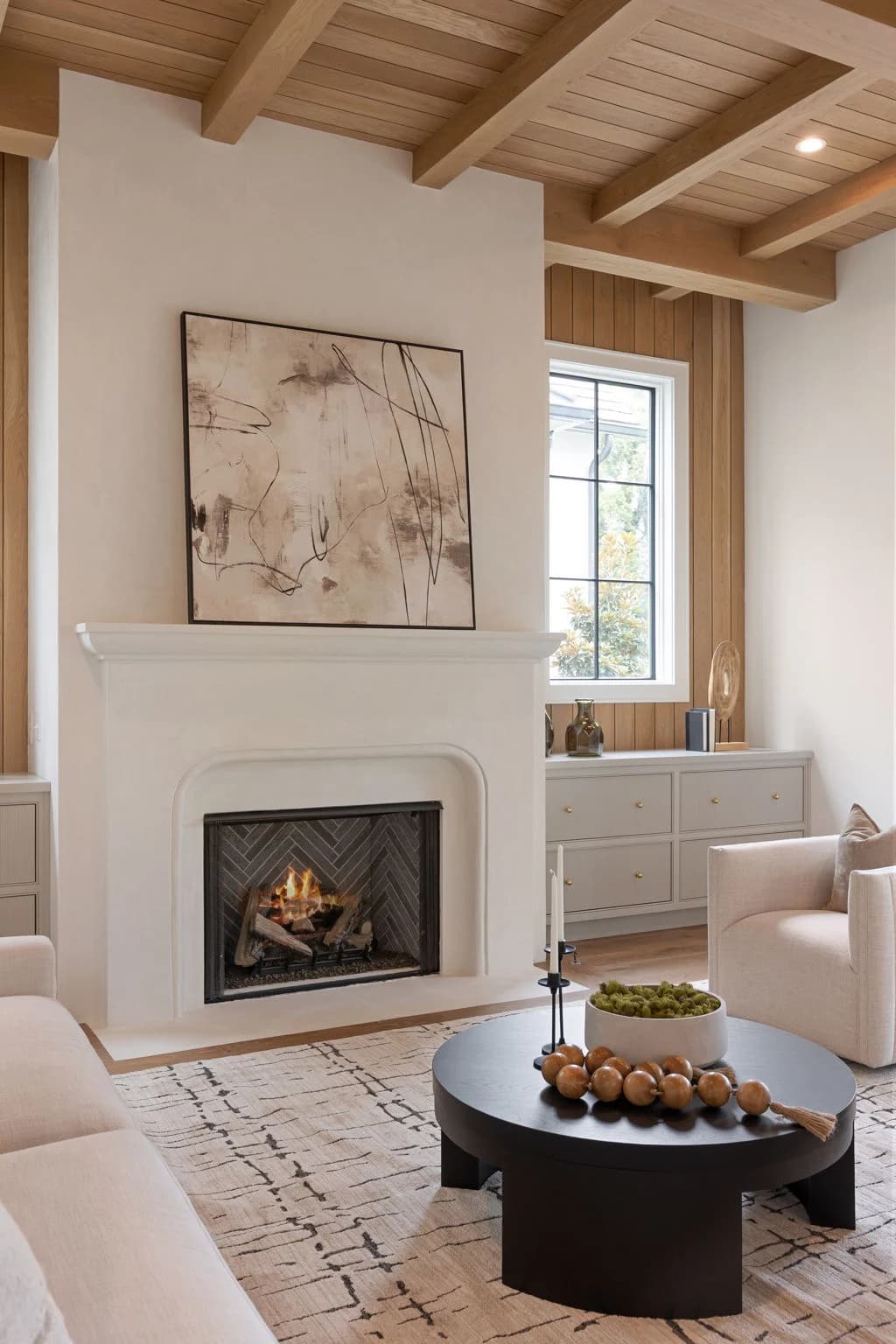 Staged living room under a tongue-and-groove wood ceiling with exposed beams, a plaster fireplace, abstract art, and a round black coffee table