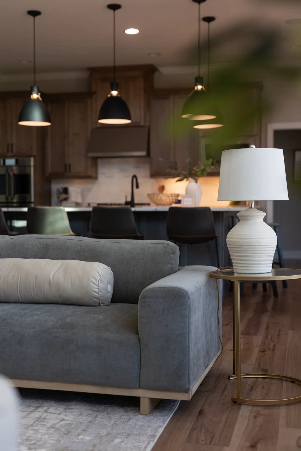 Gray sofa and lamp in a living room open to a kitchen with black pendant lights