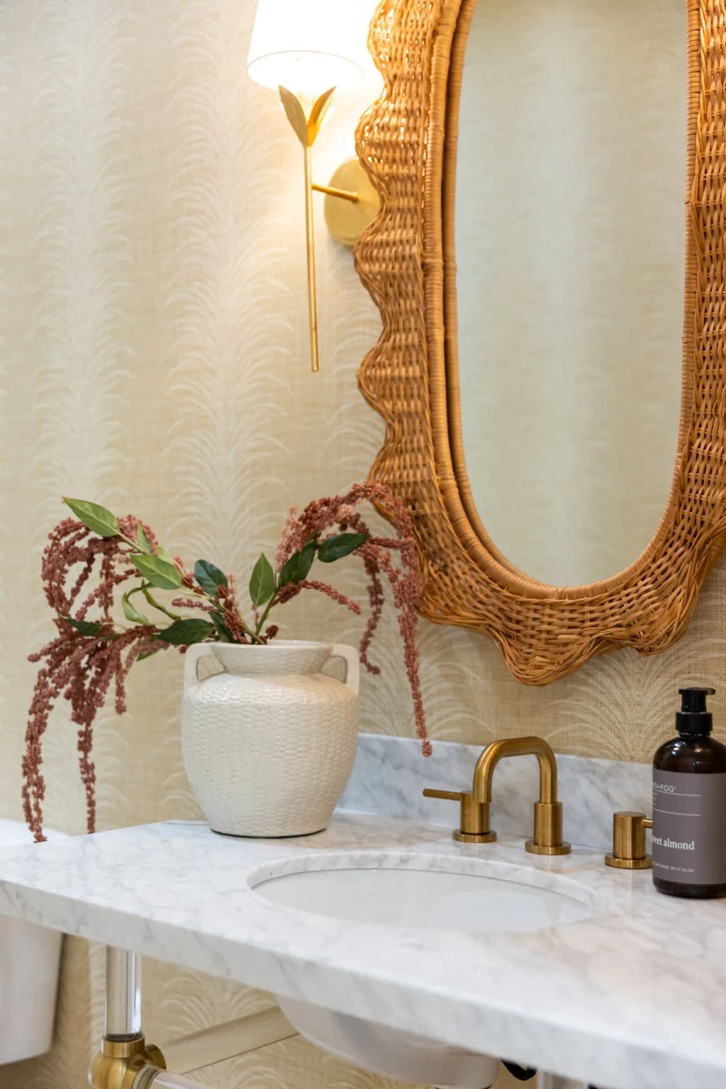 Powder room with a scalloped rattan mirror, a marble sink, a brass faucet, and a vase of trailing red blooms
