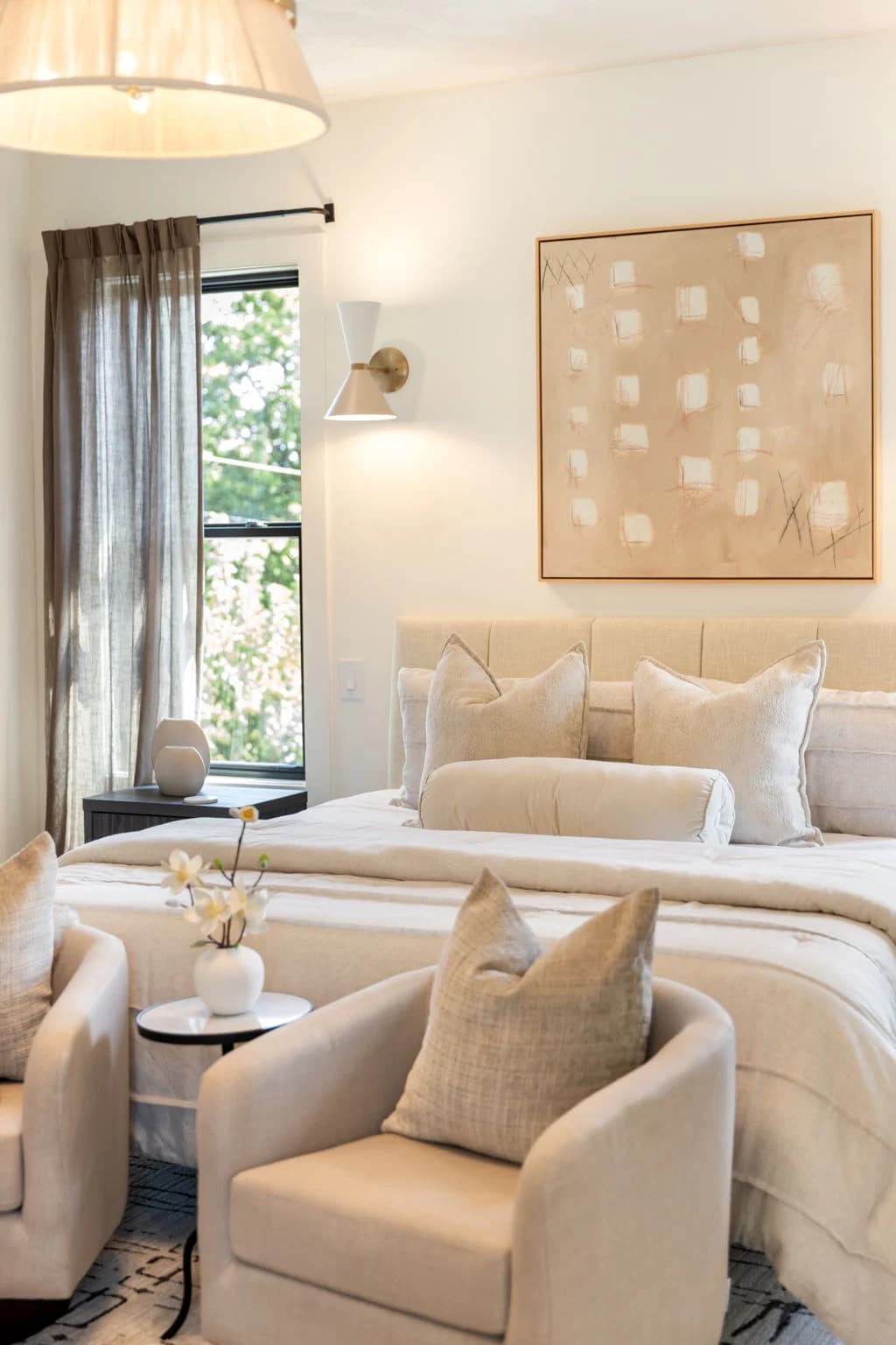 Upholstered bed with cream pillows under abstract art, a wall sconce, and a swivel chair