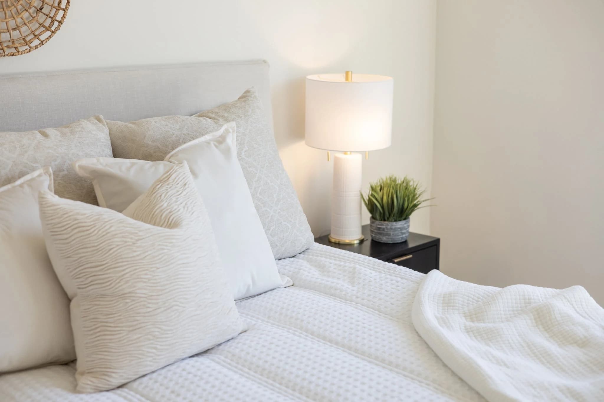 Bed layered with textured white pillows beside a white table lamp and a small plant