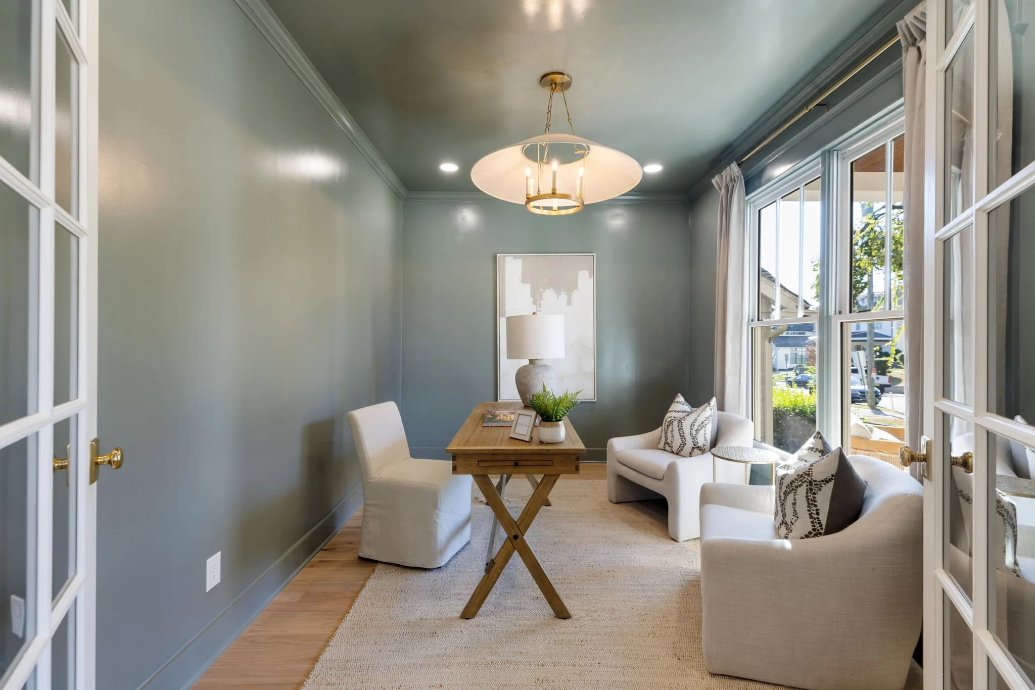 Study with glossy green gray walls, an X base wood desk, a cream sofa, and a white bowl pendant