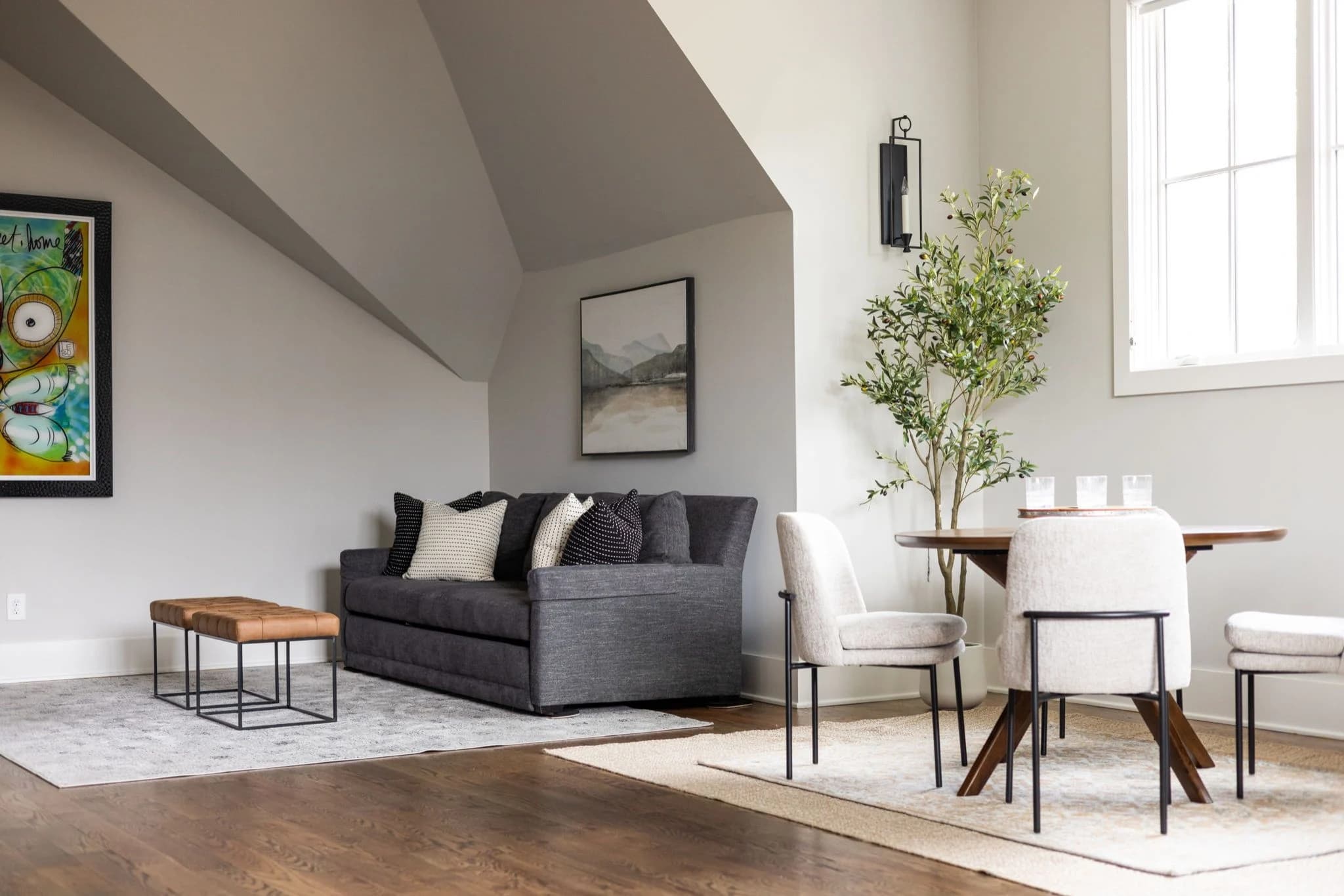 After staging: the same bonus room with a charcoal sofa, a leather bench, a dining table with cream chairs, and an olive tree