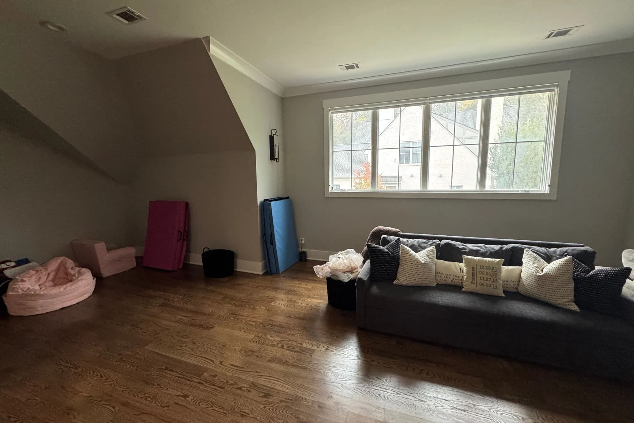 Before staging: bonus room with a dark sofa, scattered kids' play mats, and bare walls