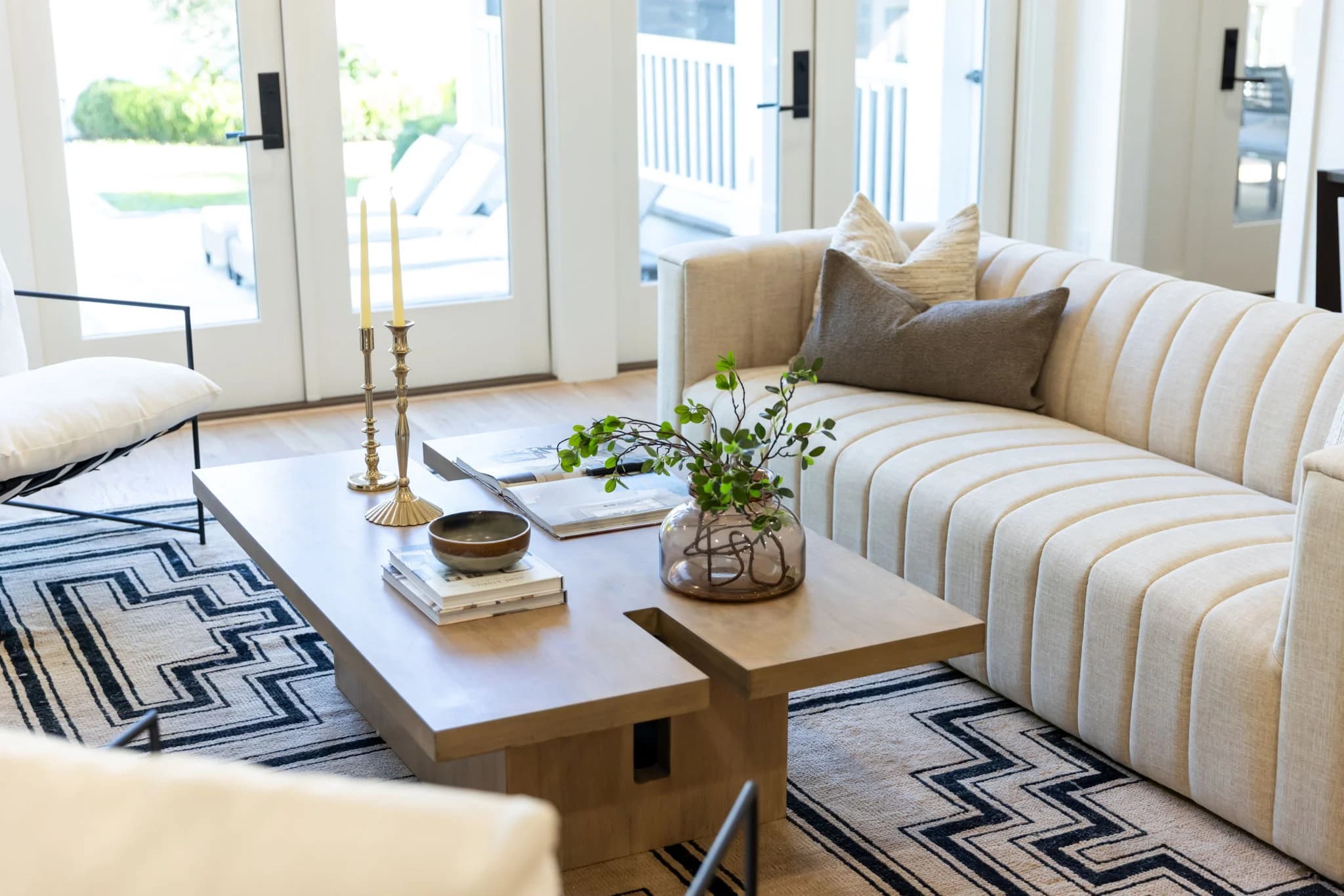 Channel tufted cream sofa and a wood coffee table with brass candlesticks on a black and cream patterned rug by French doors