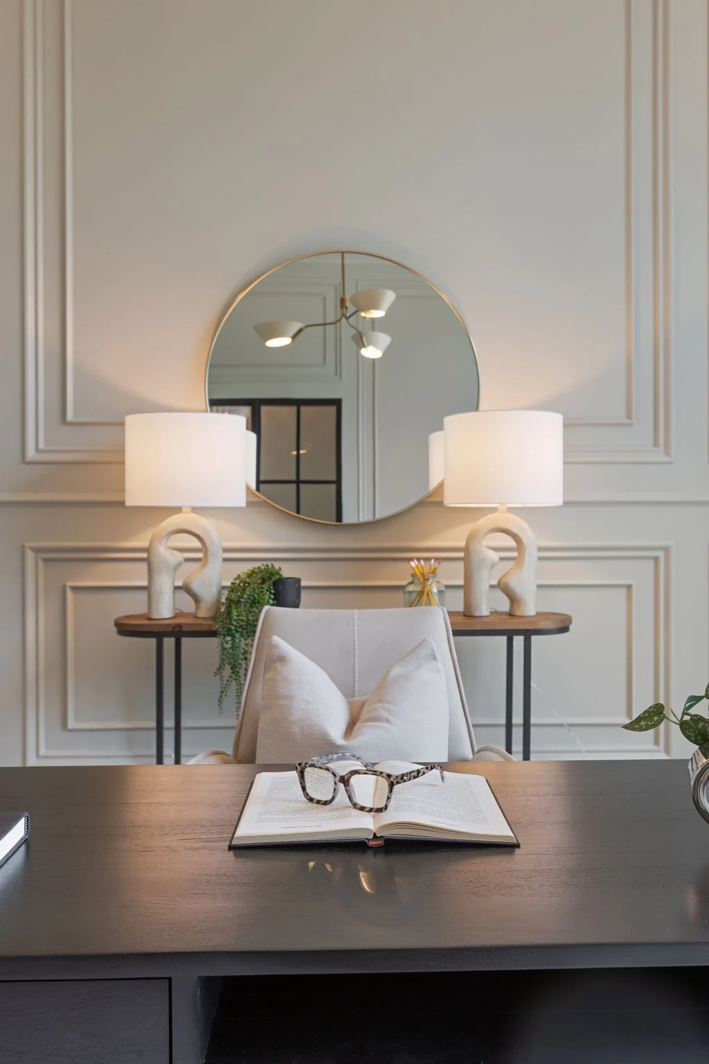 Console table styled with paired lamps, an open book, and a round mirror against a paneled wall
