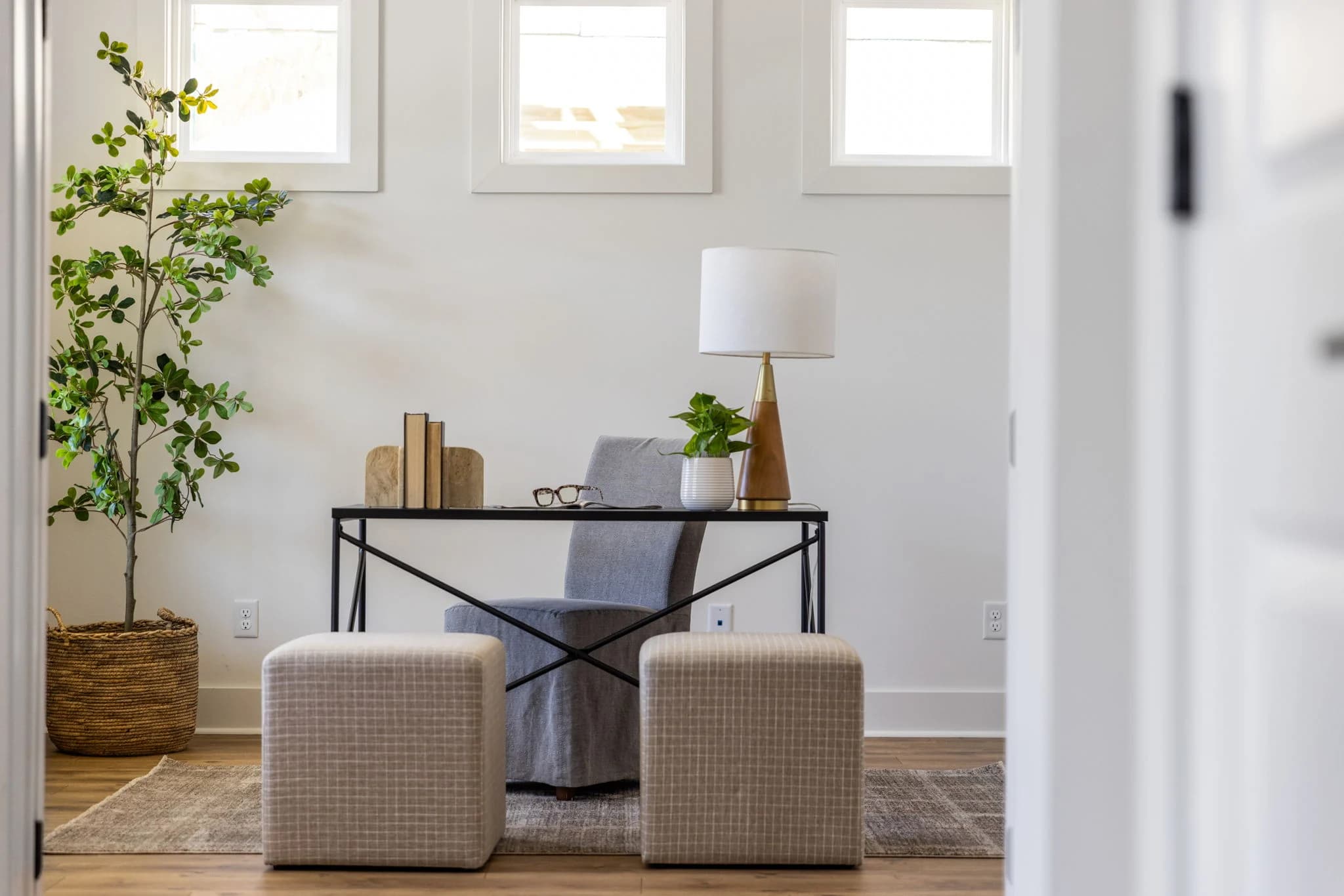 Desk nook under three small windows with a black metal desk, a slipcovered chair, two checked cube ottomans, and a potted tree