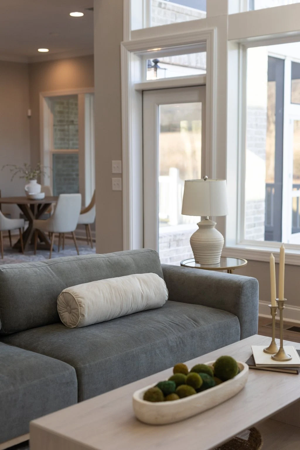 Gray sofa with a cream bolster pillow, a wood coffee table with a moss bowl, and brass candlesticks