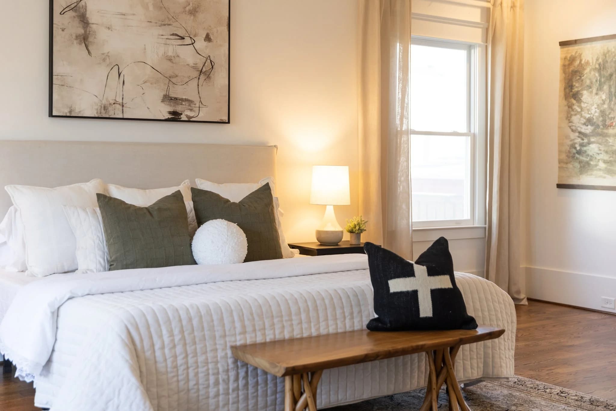 Bedroom with an olive and white bed, a black cross pillow, and a wood bench at the foot of the bed